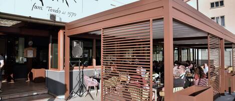 An outdoor seating area with tables and chairs, a covered section with a slatted roof, and a building with a sign that reads 'farm. Tondo dal 1970'.