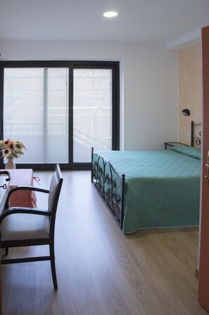 A room with a bed, a desk, and a chair. There is a window with blinds, a vase with flowers, and a wall-mounted lamp. - Ristorante Albergo Gerardo Di Masi (Caposele)