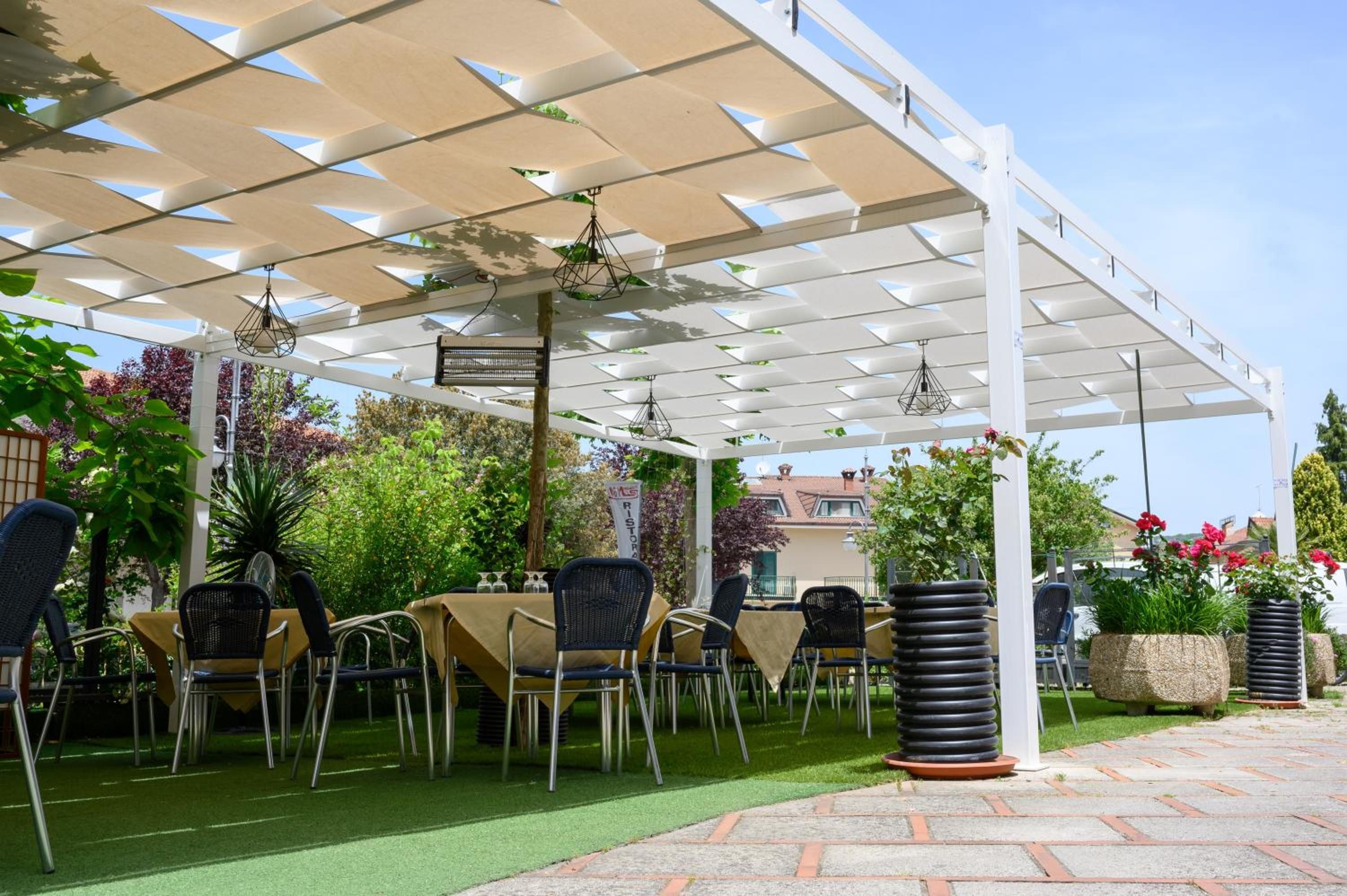 An outdoor patio with tables and chairs, covered by a white pergola, surrounded by greenery and a brick pathway.