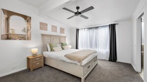 Un dormitorio con una cama, una mesita de noche, un ventilador de techo y una ventana con cortinas.