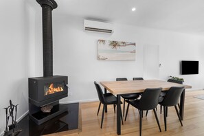 A modern living room with a wood-burning stove, a dining table with chairs, and a beach-themed painting on the wall. - Comfy Torquay Home - Pet-Friendly, Walk to Beach (Torquay)
