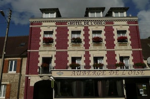 Fachada de hotel con letrero 'HOTEL DE L'OISE', varios ventanales con balcones de flores y un anuncio de 'HEINEKEN' debajo de la entrada.