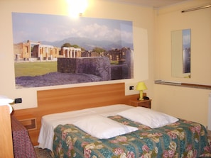 Quarto de hotel com cama, mesa de cabeceira com abajur, um quadro grande na parede e espelho.