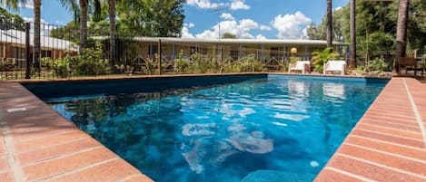 Piscina con borde de ladrillo, rodeada de palmeras y una zona cubierta con sillones de descanso.