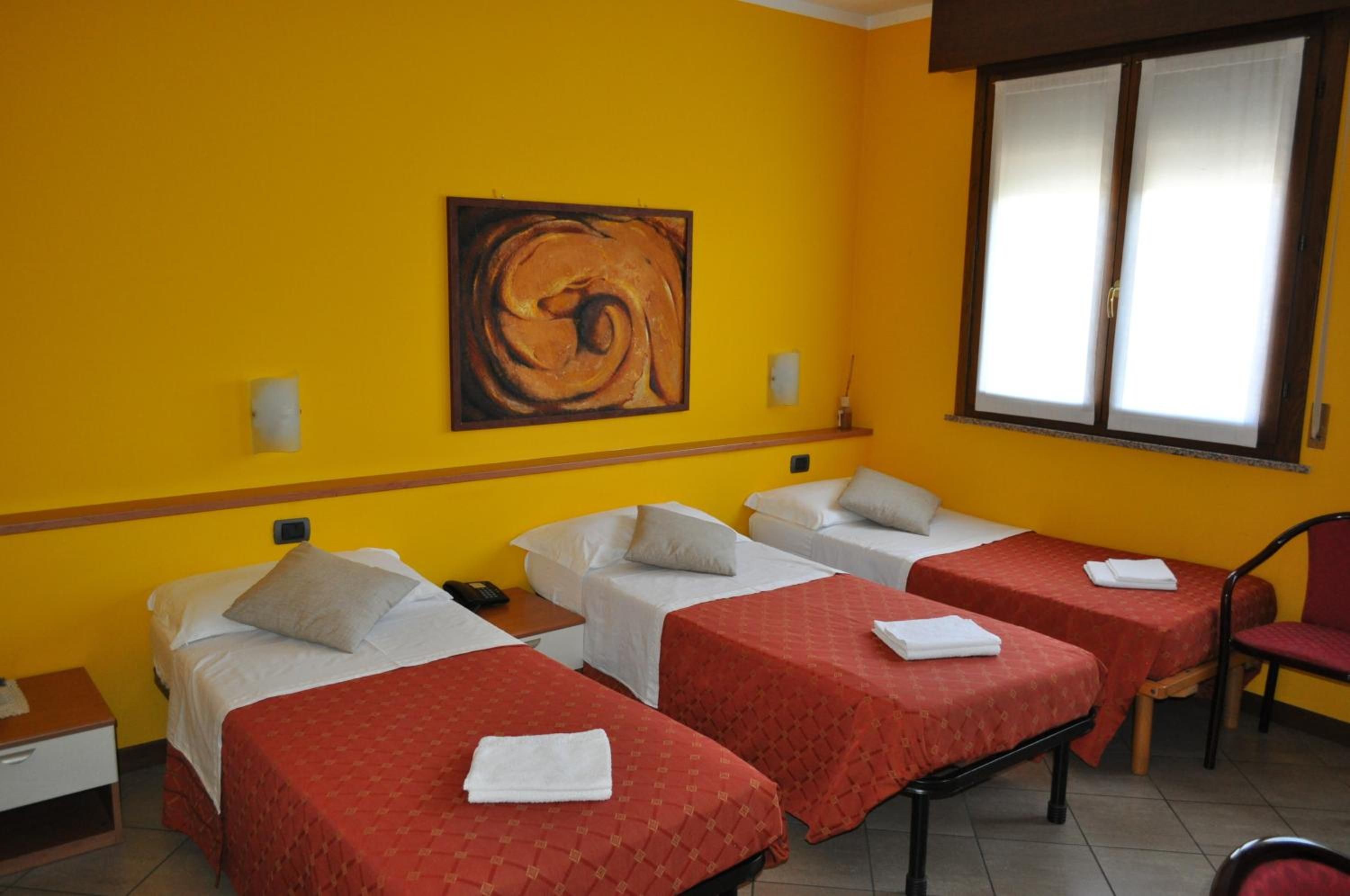 Une chambre d’hôtel avec trois lits simples, une chaise rouge, un tableau au mur et une fenêtre avec des stores.