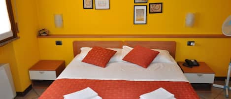 A hotel room with a bed, two bedside tables, a nightstand, a wall-mounted fan, and artwork on the wall.