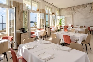 Un restaurante con varias mesas, cada una con manteles blancos, platos y cubiertos. Amplias ventanas ofrecen vista al mar.
