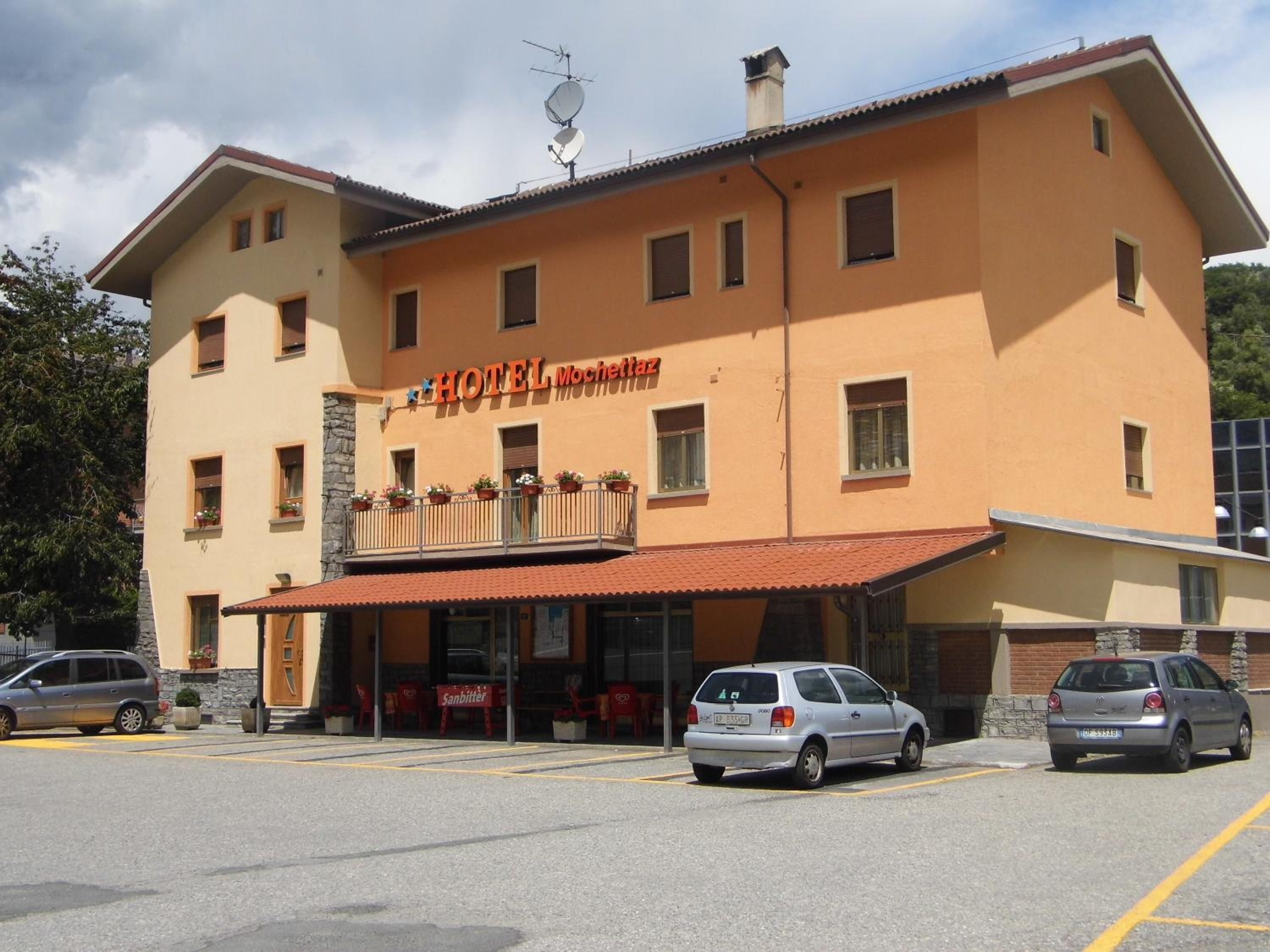 Un hotel de dos pisos con techo rojo y un cartel que dice 'HOTEL Mochetaz'. Hay dos coches estacionados al frente.