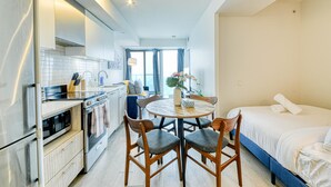Et lille hotelværelse med tekøkken, et rundt spisebord med fire stole, en enkelt seng og et stort vindue, der giver naturligt lys.