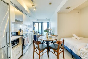 Quarto de hotel compacto com cozinha compacta, uma mesa de jantar redonda com quatro cadeiras, uma cama de solteiro e uma grande janela que oferece iluminação natural.