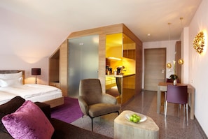 Ett modernt hotellrum med en säng, en sittgrupp med en lila kudde, ett litet bord med en skål med frukt, en stol och en liten köksdel.
