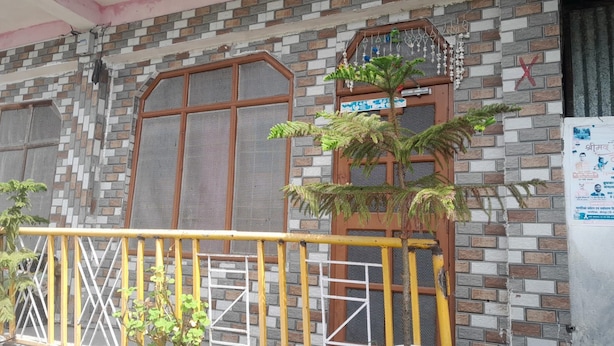 Fachada de edificio con patrón de ladrillo, dos ventanas grandes con rejas metálicas, un árbol pequeño y una barandilla amarilla.