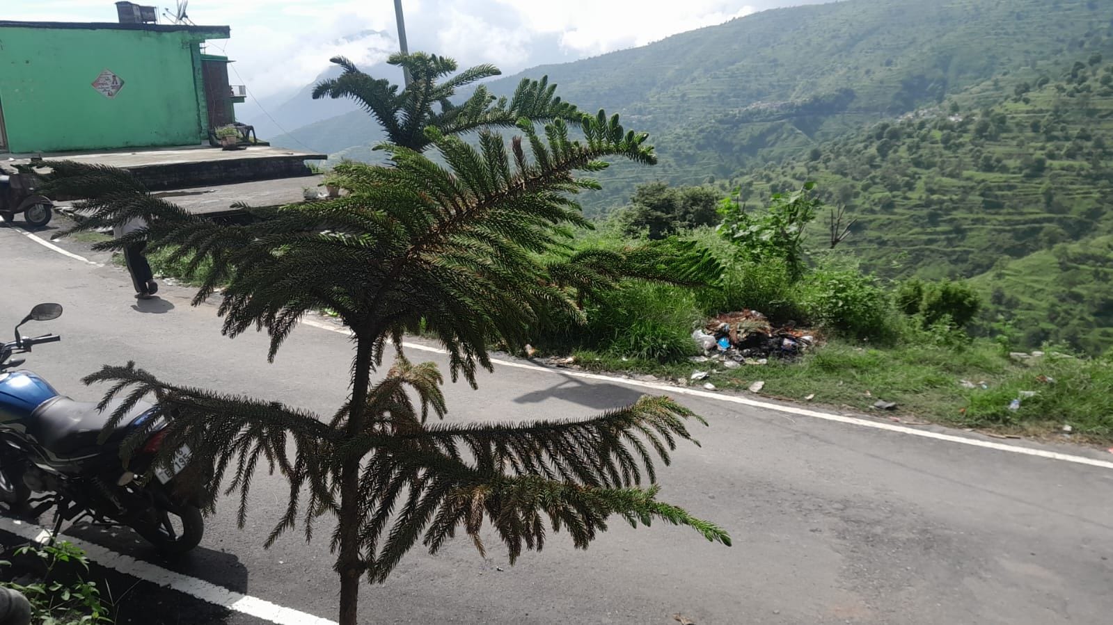 A road with a motorcycle parked on the side, surrounded by greenery and a hilly landscape in the background.