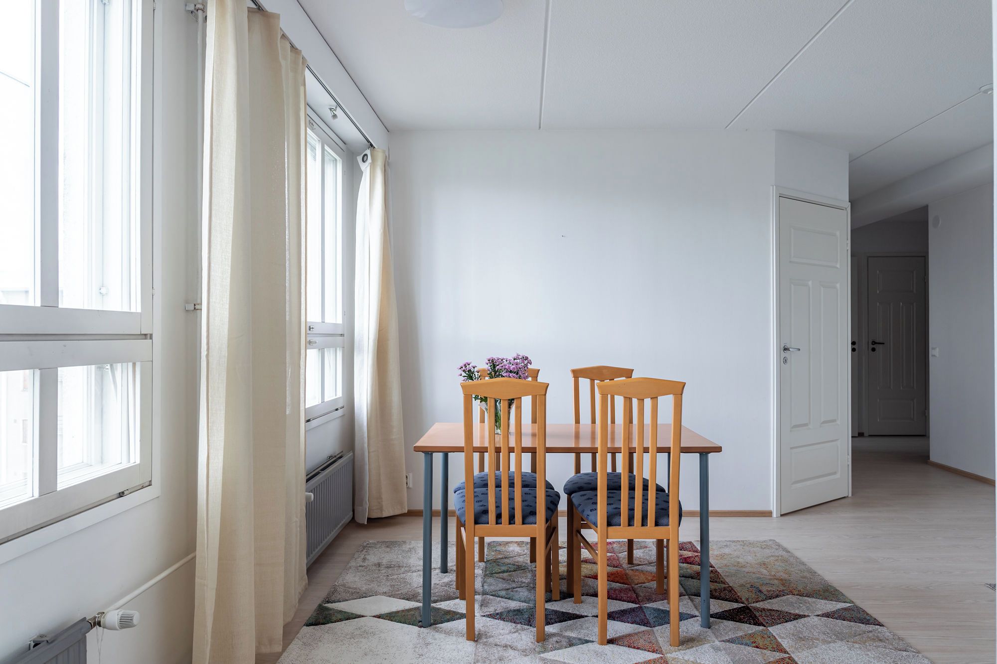 Una habitación con una mesa de comedor, cuatro sillas, un radiador, una ventana con cortinas y una puerta que da a otra habitación.