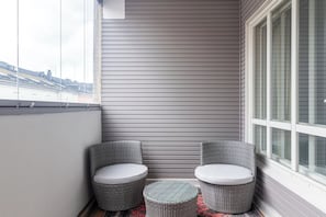 Un balcón en esquina con dos sillas de mimbre y una pequeña mesa redonda, una ventana con persianas, y vistas a edificios al fondo.