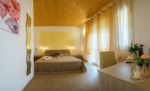A hotel room with a wooden ceiling, a bed with patterned bedding, a desk with a vase and a remote control, and a window with curtains.