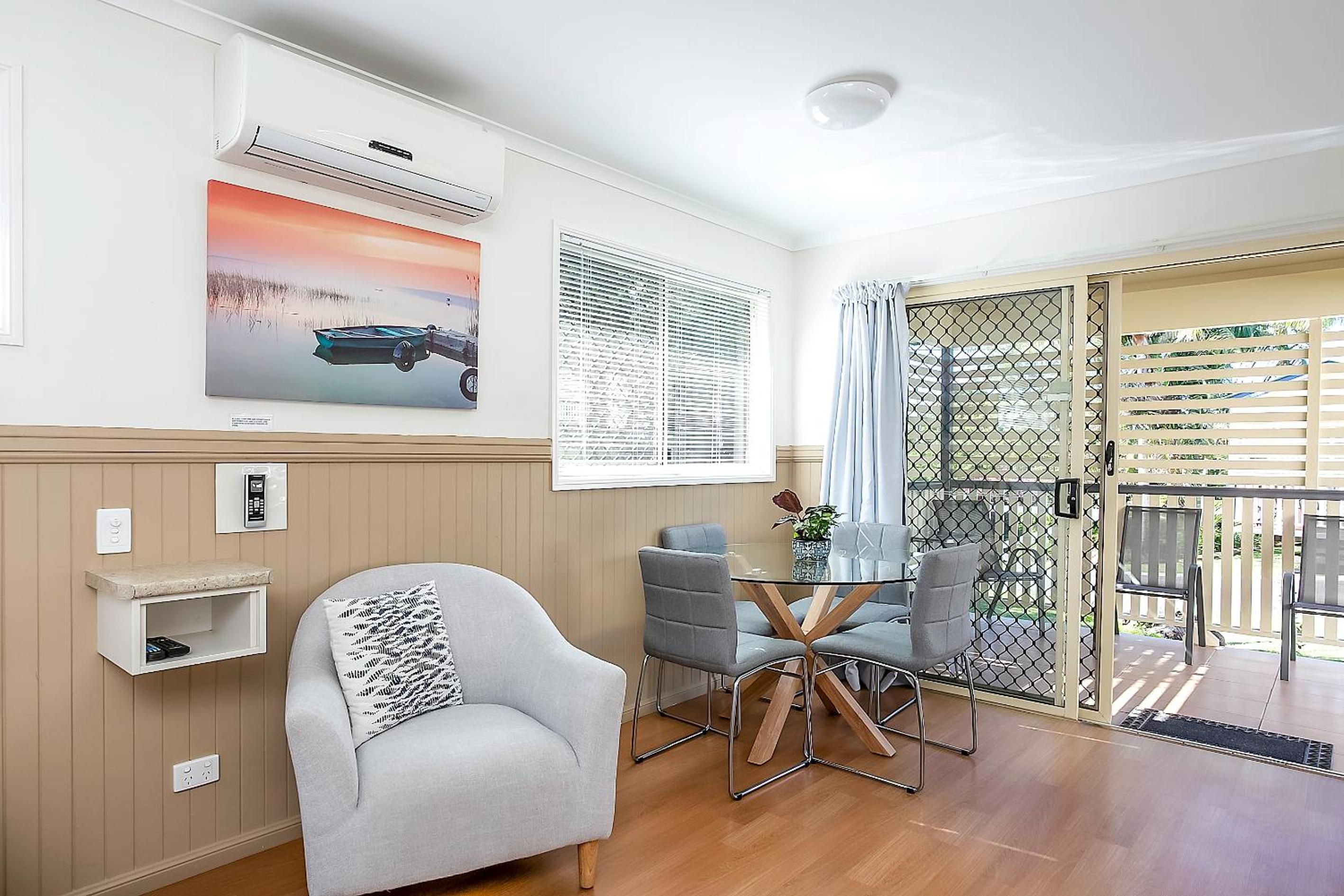 A room with a single grey armchair, a small table with chairs, a wall-mounted air conditioner, a window with blinds, and a door leading outside.