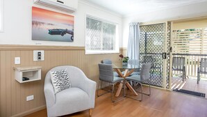 A room with a single grey armchair, a small table with chairs, a wall-mounted air conditioner, a window with blinds, and a door leading outside.