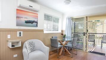 A room with a single grey armchair, a small table with chairs, a wall-mounted air conditioner, a window with blinds, and a door leading outside.