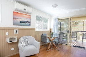 A room with a single grey armchair, a small table with chairs, a wall-mounted air conditioner, a window with blinds, and a door leading outside.