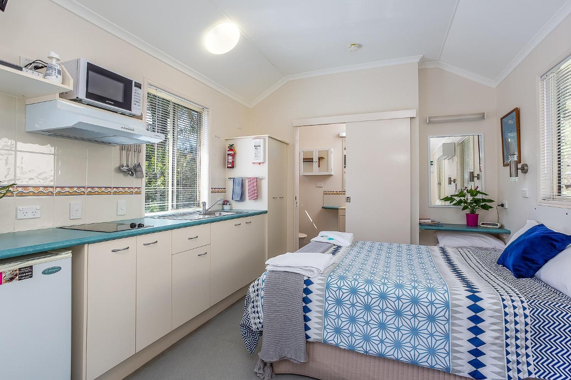 A compact hotel room with a kitchenette, a bed with patterned bedding, a bedside table with a plant, and a mirror on the wall.