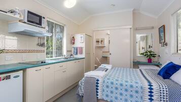 A compact hotel room with a kitchenette, a bed with patterned bedding, a bedside table with a plant, and a mirror on the wall.