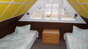 A small, triangular attic bedroom with two beds, a wooden chest of drawers, and a window with a view of the outside.