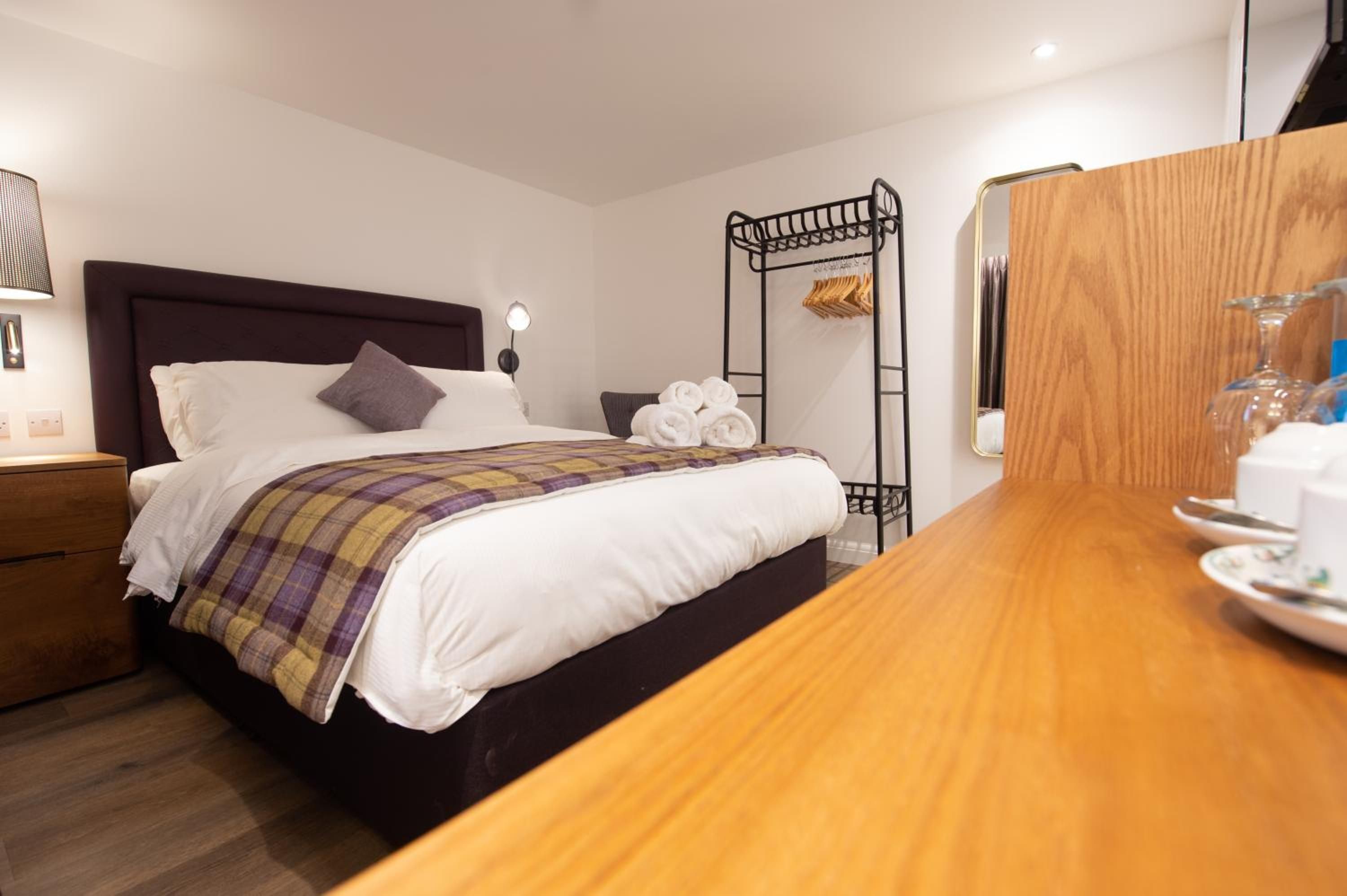 A hotel room with a bed, a wooden nightstand, a bedside table, a hanging towel rack, and a wooden dresser.