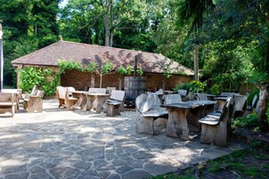 Un patio exterior con suelo de piedra y mobiliario de madera rústica, rodeado de vegetación y un edificio de ladrillo.