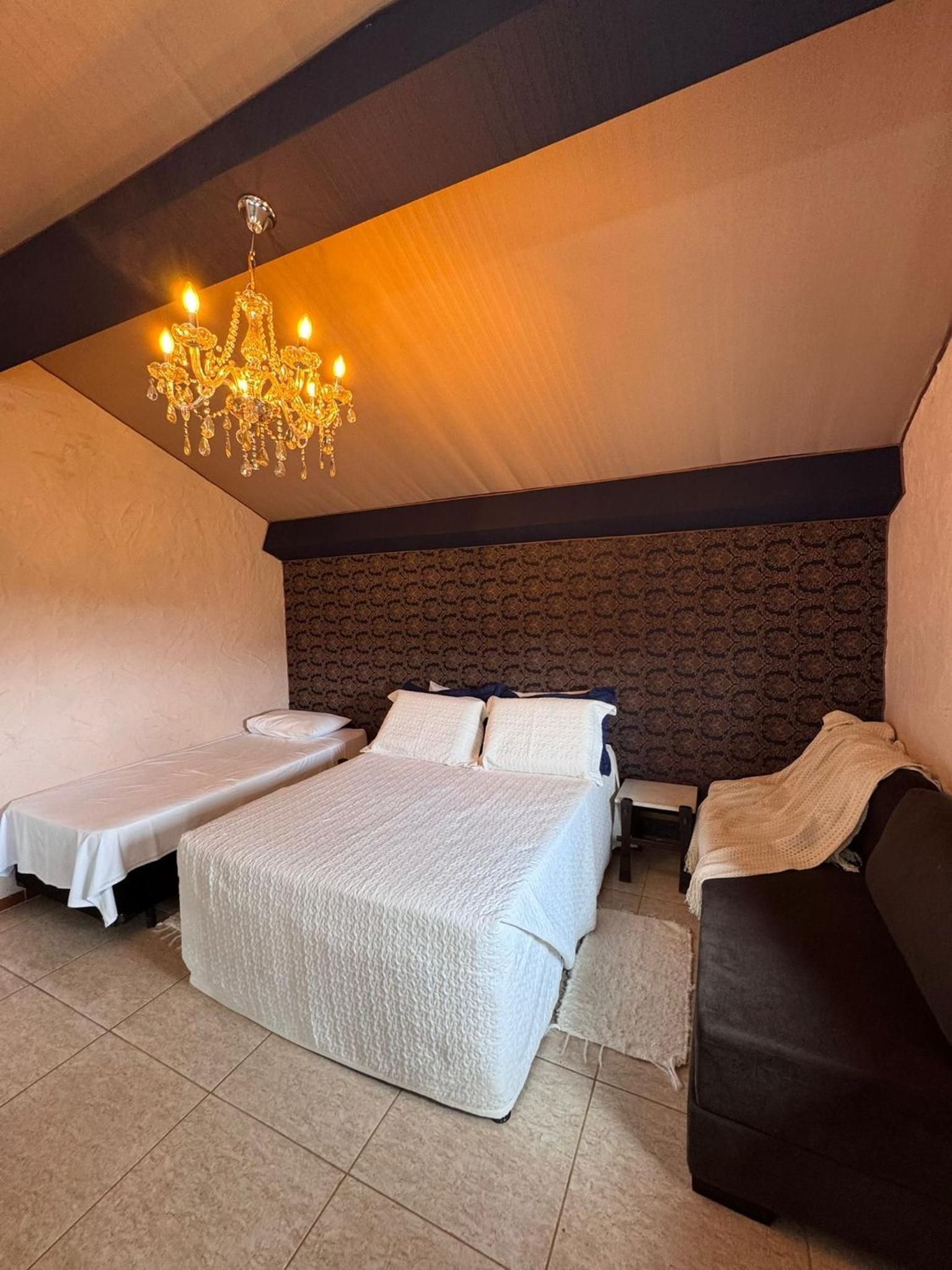 Quarto aconchegante com uma cama grande, duas camas menores, cabeceira escura e um candelabro.