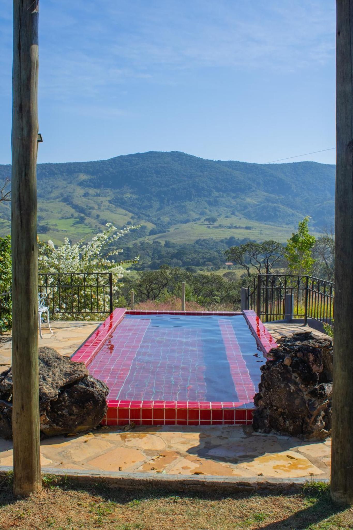 Piscina com deck ladrilhado em vermelho e azul, rodeada por uma cerca de metal, com colinas suaves e céu claro ao fundo.