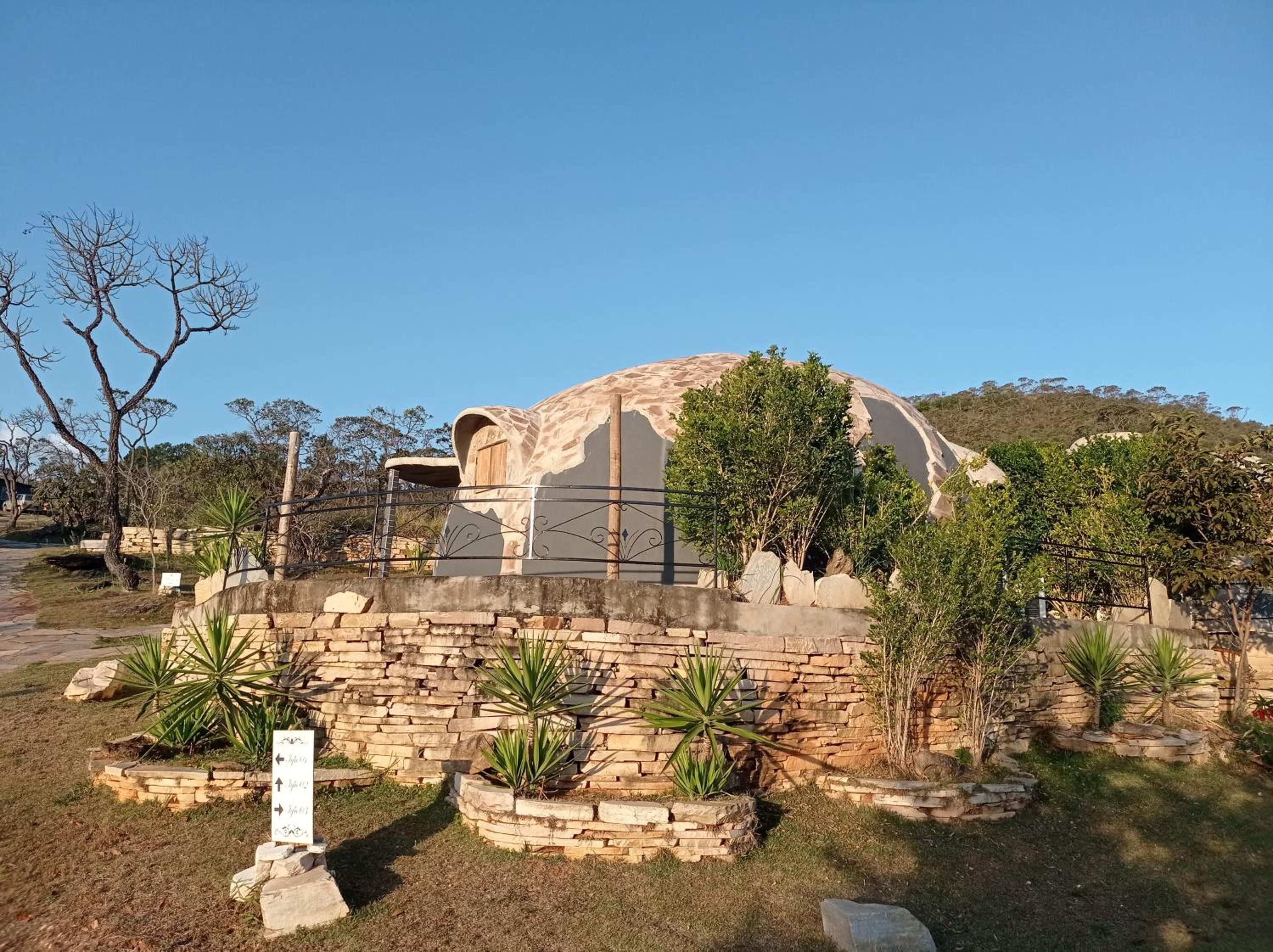 Um edifício em formato de cúpula com um muro de pedra à frente, rodeado por diversas plantas e um céu claro.