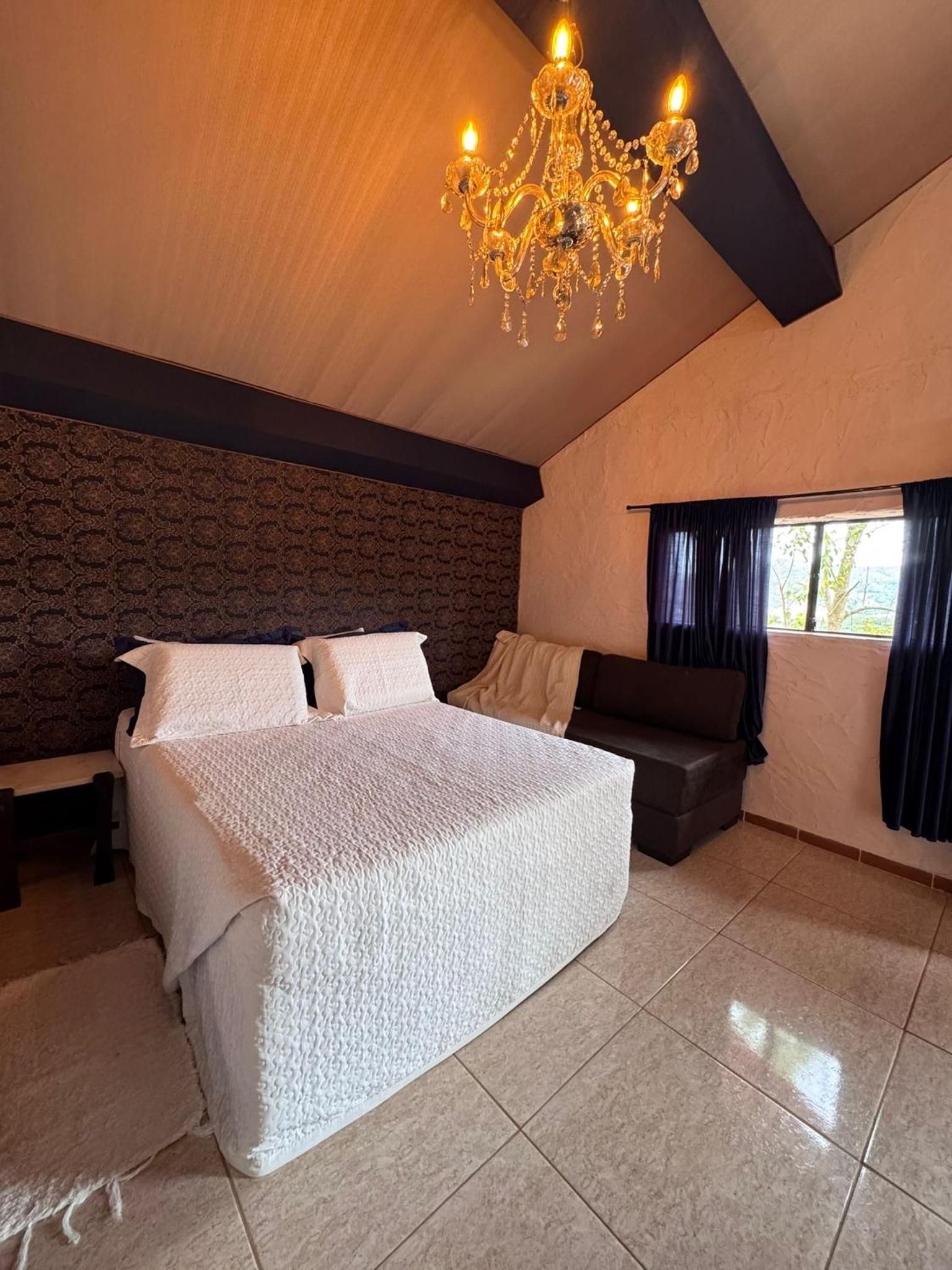 Quarto com cama grande, sofá escuro, candelabro e janela com cortinas.