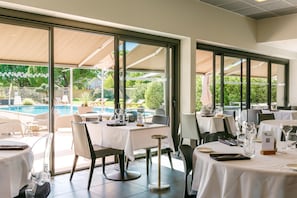 An outdoor dining area with tables and chairs, large windows, and a view of a pool and greenery. - Sous Les Pins (Etoile-sur-Rhone)