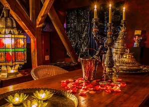 Um quarto com pouca luz, com uma mesa de madeira, uma luminária de vitrais, um candelabro e um jarro de metal decorativo.