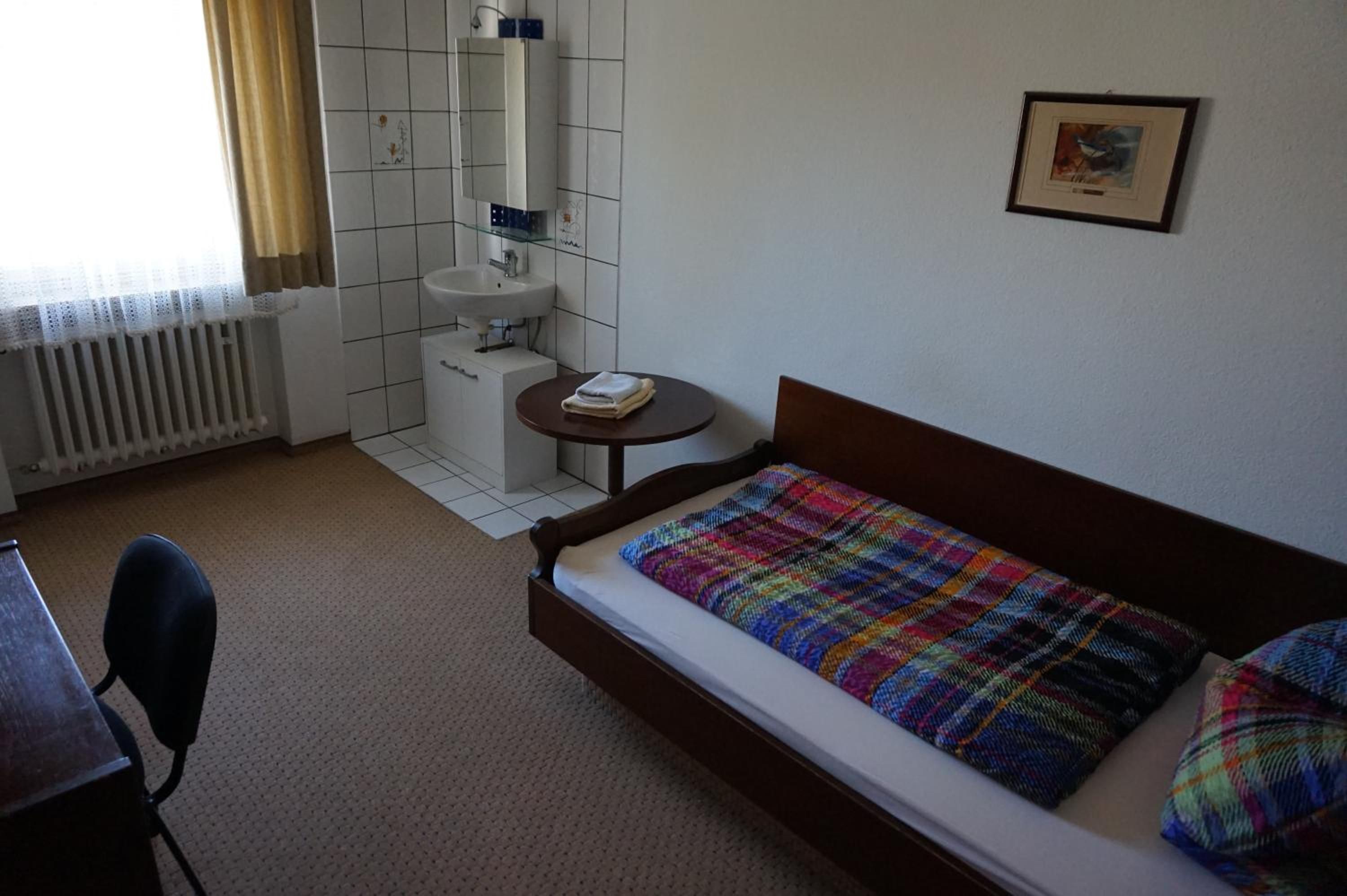 A bedroom with a bed, a small bedside table, a chair, a radiator, a window with curtains, and a wall-mounted picture.