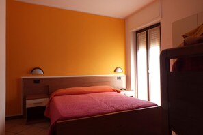 A bedroom with an orange wall, a bed with a red bedspread, a nightstand, a desk, and a window with blinds. - Albergo Ristorante Innocenti (Ardenno)