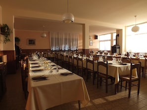 A restaurant with long tables set for dining, wooden chairs, and hanging pendant lights. - Albergo Ristorante Innocenti (Ardenno)