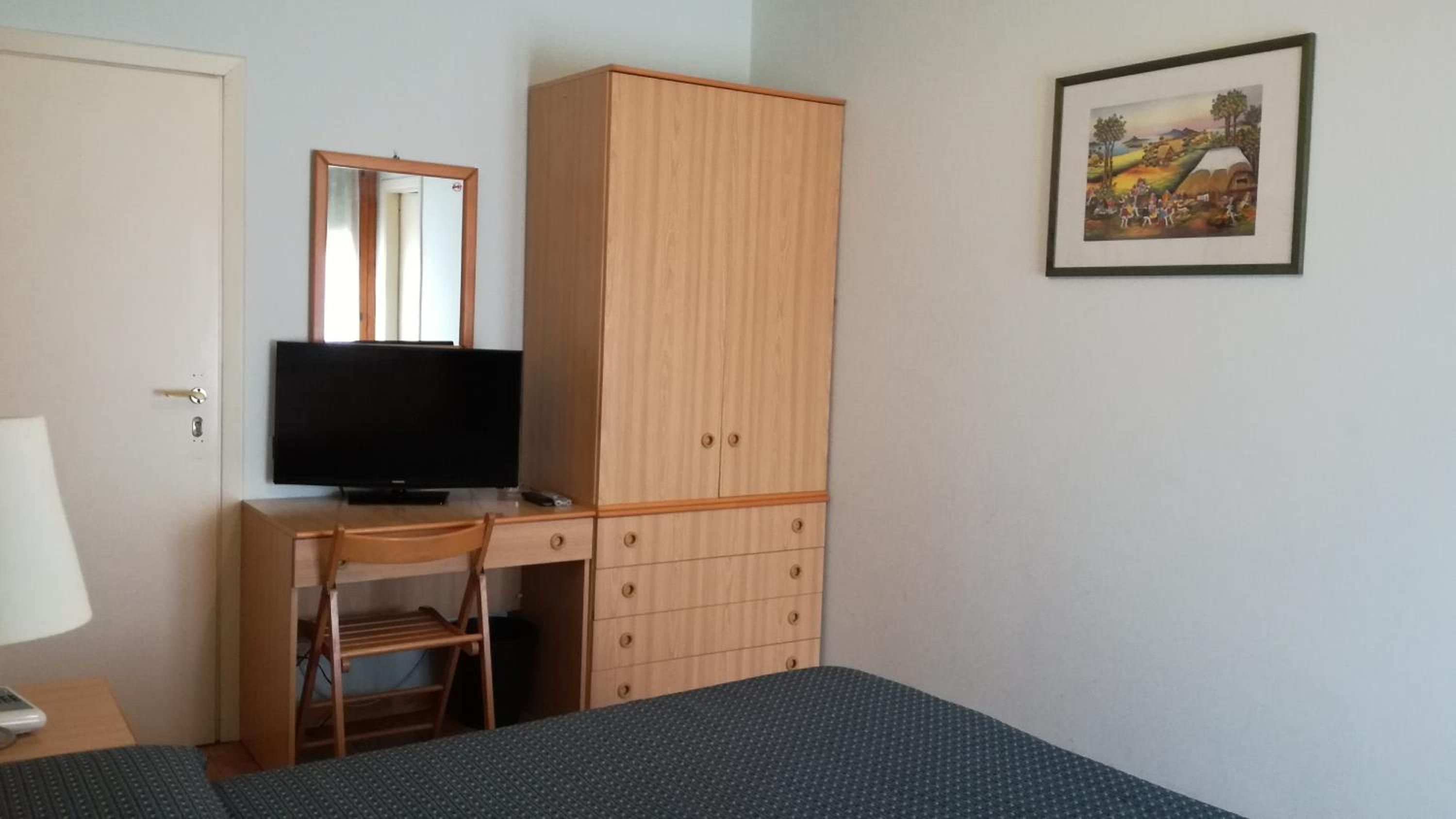 A bedroom with a bed, a desk with a television, a wardrobe, and a framed picture on the wall.