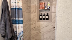 Área de duche com cortina de chuveiro estampada, prateleira com artigos de higiene e chão em azulejo.