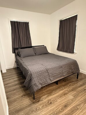 A single bed with a dark-colored headboard and footboard, positioned against a wall with two windows, each with a dark curtain. The floor is wooden with a herringbone pattern.