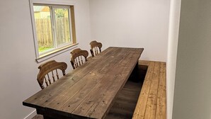 Um quarto com uma mesa de madeira comprida, quatro cadeiras, um banco, uma janela e uma luminária no teto.