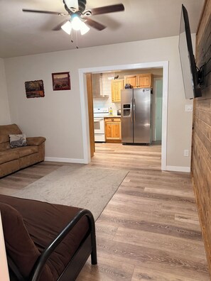 A modern living room with a brown sofa, a bed, and a ceiling fan. A kitchen with wooden cabinets and a refrigerator is visible through an open doorway. - Beautiful 2-bedroom, 2-bathroom house with a yard in beautiful Halfway Oregon. (Halfway)