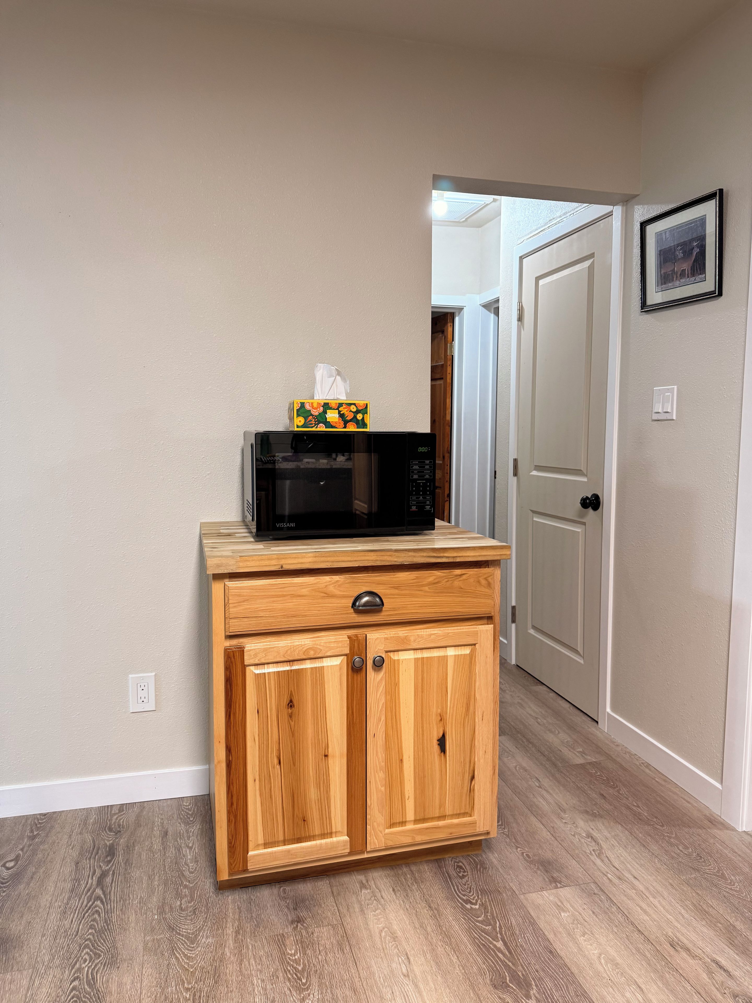 A kitchen area with a microwave on a wooden cabinet, a door leading to another room, and a wall-mounted light switch.