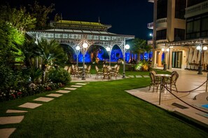 Uma área de estar externa com mesas e cadeiras, cercada por um gramado bem cuidado e iluminação decorativa.