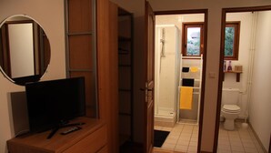 Habitación con televisión sobre un aparador de madera, un espejo y una puerta abierta que da a un baño con ducha, inodoro y estante con artículos de aseo.