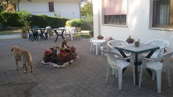 Patio esterno con tavolo e sedie, circondato da piante in vaso e un cane.