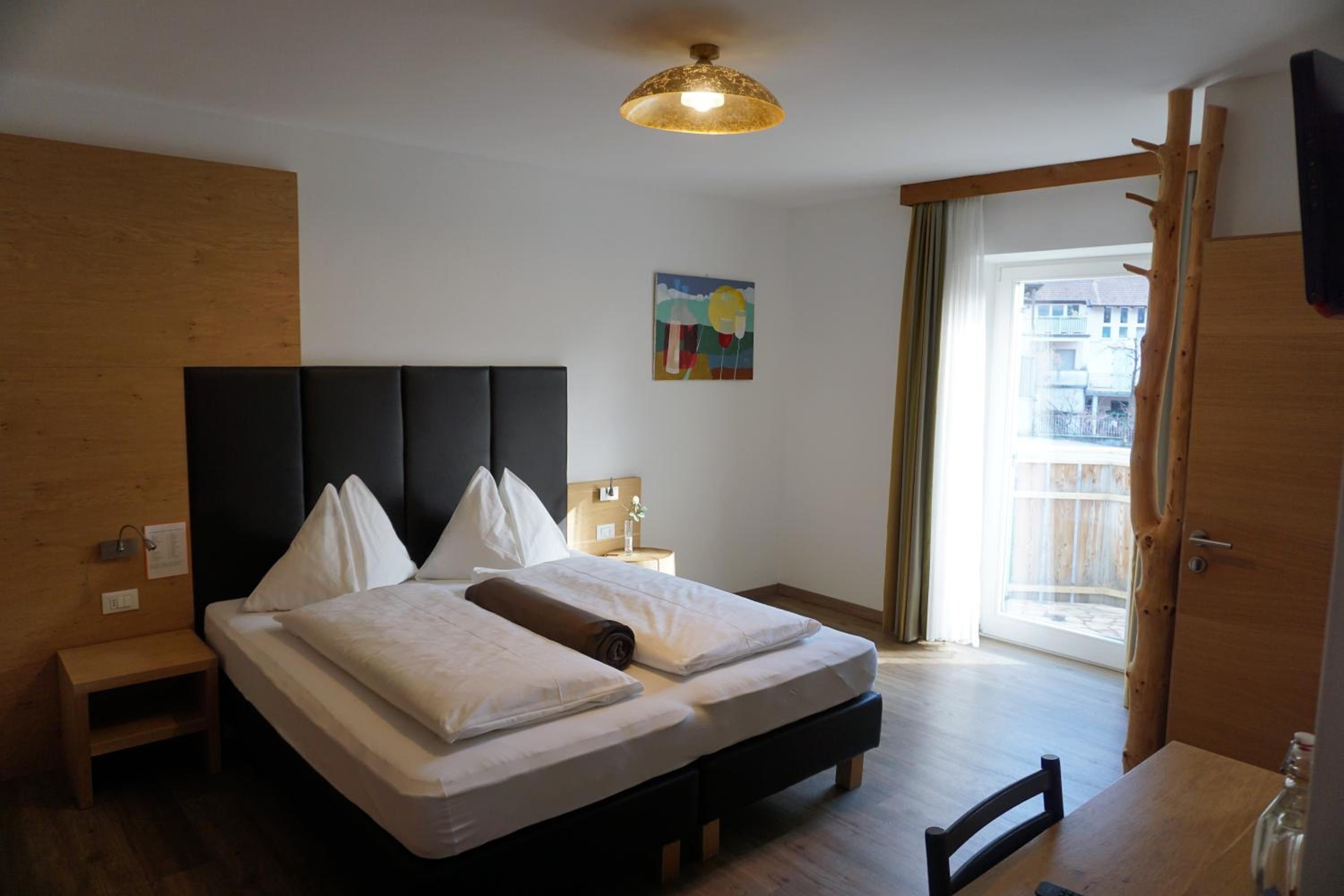 A hotel room with a large bed, a bedside table, a nightstand, a painting on the wall, and a view through a door.