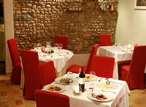 A restaurant with stone walls and red chairs. Tables are set with plates, glasses, and a bottle. - Hotel Le Macine (Vittorio Veneto)