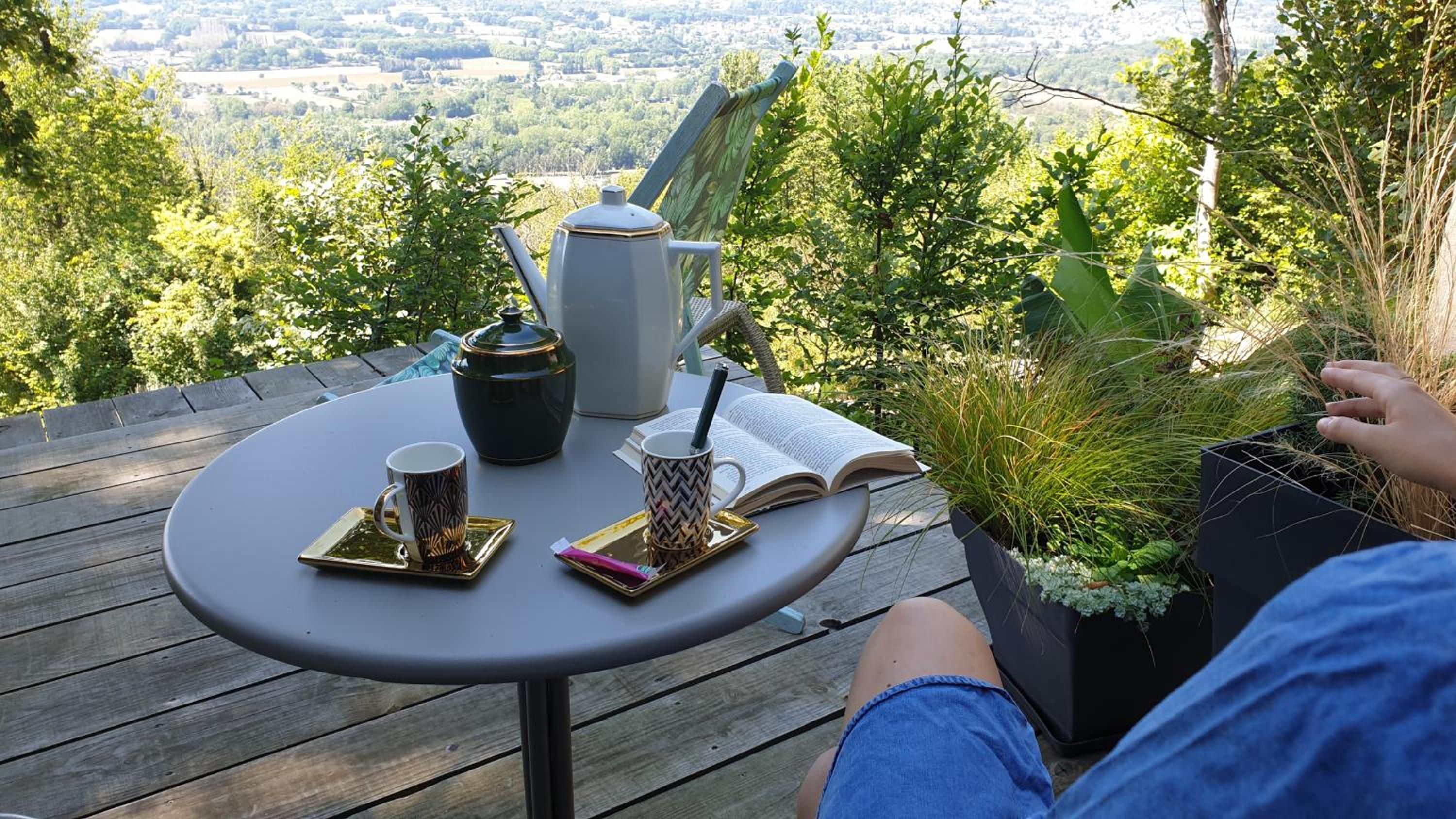 A round table with a book, a mug, and a teapot on a deck overlooking a landscape.